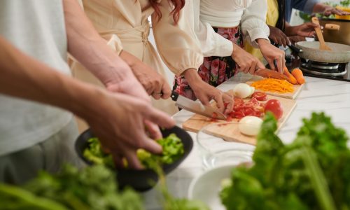 Row of young intercultural friends with sharp knives chopping fresh vegetables for salad while standing by table and cooking dinner
